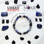 A beaded bracelet with blue, gray, black, and silver beads, displayed on a white marble surface surrounded by black and blue crystals. Text overlays read 'A Collaboration with URBAN INVESTIGATOR + MOXIE MALAS' and 'Abandonment Issues'.