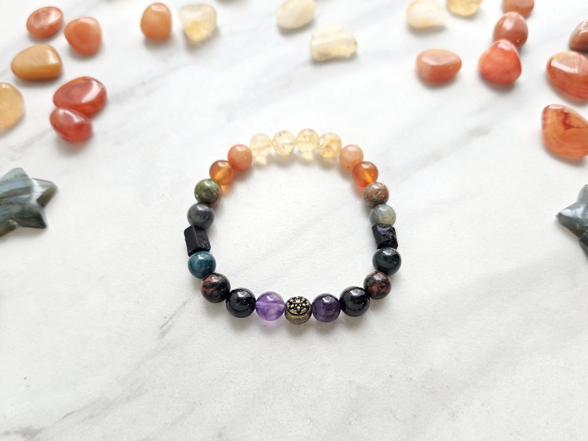A beaded stretch bracelet displayed on a white marble surface, surrounded by scattered polished stones. The bracelet features a gradient of gemstone beads from orange and yellow at the top, through green and gray, to dark purple and black, with a central metallic gold and silver patterned bead.