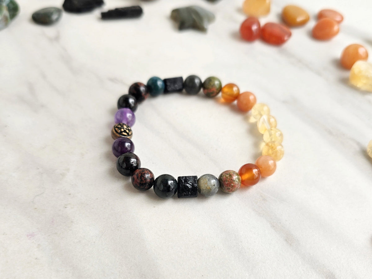 A beaded stretch bracelet featuring a gradient of gemstone beads from dark black and purple at one end to translucent orange and yellow at the other, displayed on a light marble surface with other stones in the background.