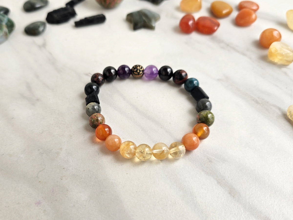 A multicolor beaded chakra bracelet displayed on a light marble surface, surrounded by loose natural stones.