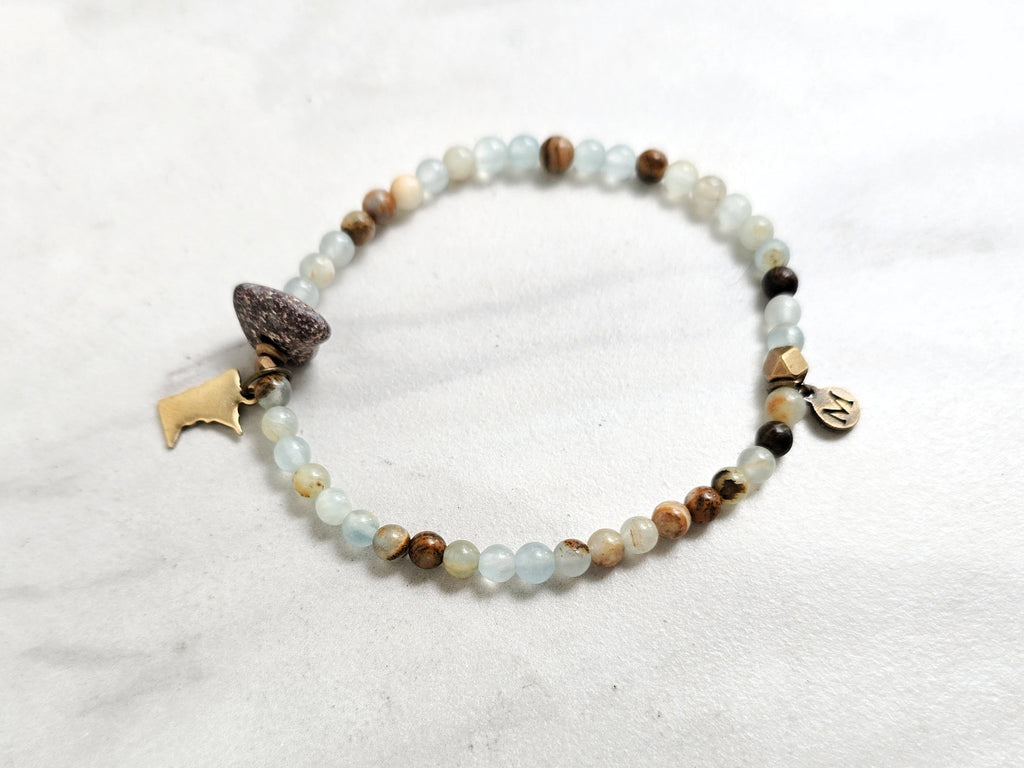 A beaded stretch bracelet featuring light blue-green aquatine calcite beads, brown and cream accent beads, a dark brown bead with a white inlay, a small gold-toned charm, and a gold-toned Minnesota state silhouette charm, displayed on a white marble background with text overlays. MN State Fair Exclusive Moxie Malas