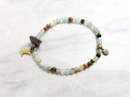 A beaded stretch bracelet featuring light blue-green aquatine calcite beads, brown and cream accent beads, a dark brown bead with a white inlay, a small gold-toned charm, and a gold-toned Minnesota state silhouette charm, displayed on a white marble background with text overlays. MN State Fair Exclusive Moxie Malas