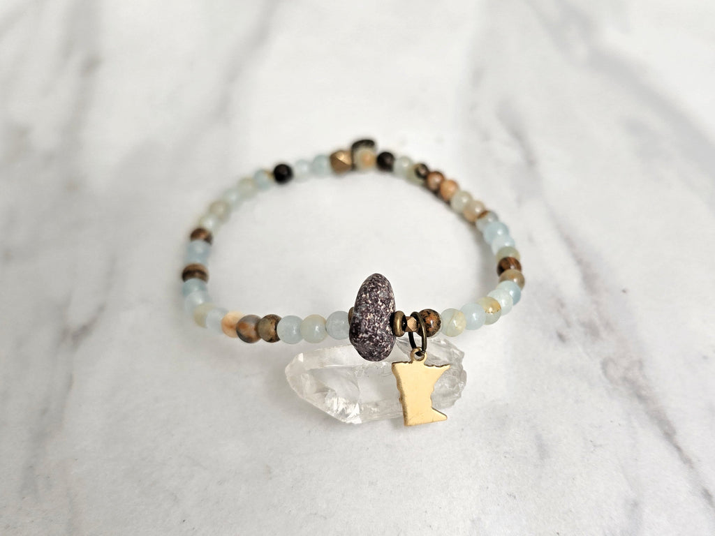 A beaded stretch bracelet featuring light blue-green aquatine calcite beads, brown and cream accent beads, a dark brown bead with a white inlay, a small gold-toned charm, and a gold-toned Minnesota state silhouette charm, displayed on a white marble background with text overlays. MN State Fair Exclusive Moxie Malas