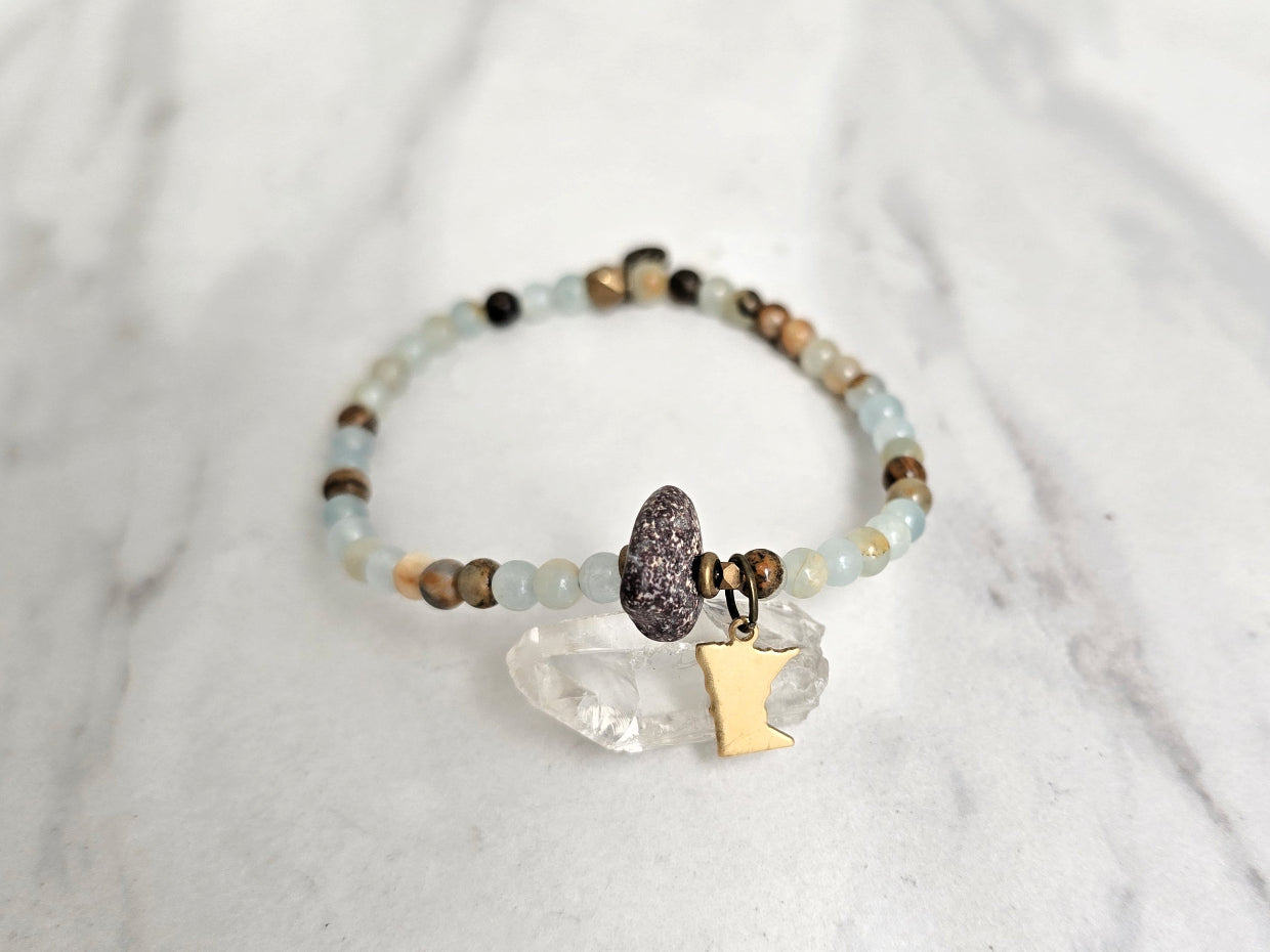 A beaded stretch bracelet featuring light blue-green aquatine calcite beads, brown and cream accent beads, a dark brown bead with a white inlay, a small gold-toned charm, and a gold-toned Minnesota state silhouette charm, displayed on a white marble background with text overlays. MN State Fair Exclusive Moxie Malas