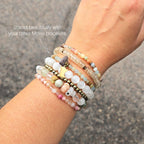 A beaded stretch bracelet featuring light blue-green aquatine calcite beads, brown and cream accent beads, a dark brown bead with a white inlay, a small gold-toned charm, and a gold-toned Minnesota state silhouette charm, displayed on a white marble background with text overlays. MN State Fair Exclusive Moxie Malas