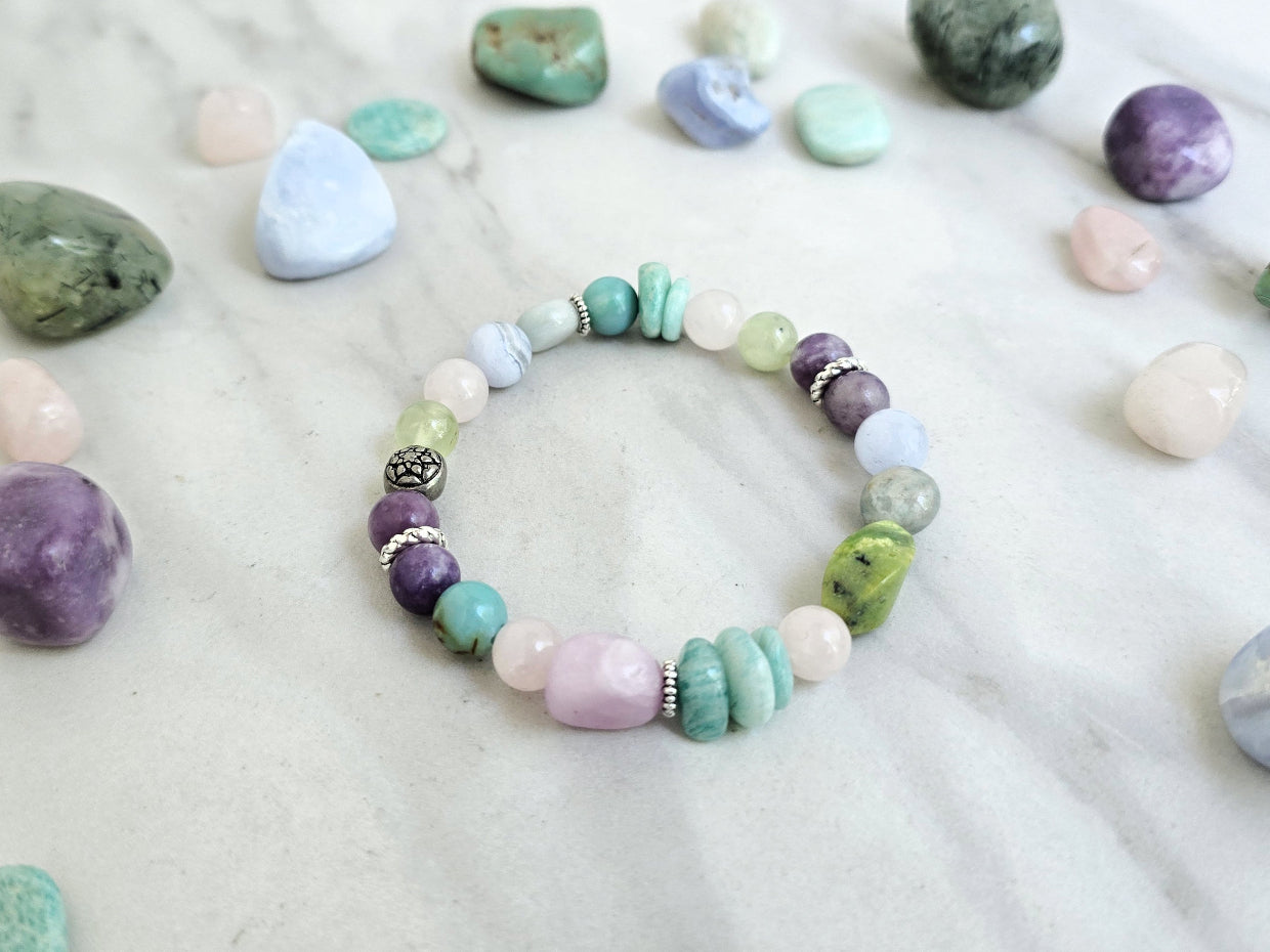 A beaded bracelet with pastel and earthy tones is arranged on a light marble surface, surrounded by polished crystals. Text overlays read 'MOXIE Monthly', 'Joyful Service', and 'September 2026'.