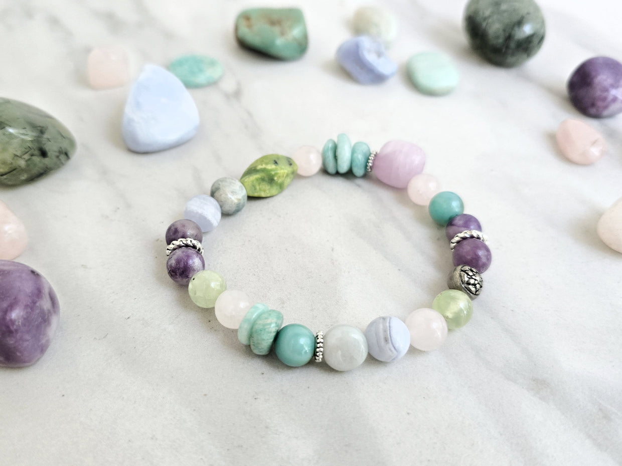 A beaded bracelet with pastel and earthy tones is arranged on a light marble surface, surrounded by polished crystals. Text overlays read 'MOXIE Monthly', 'Joyful Service', and 'September 2026'.