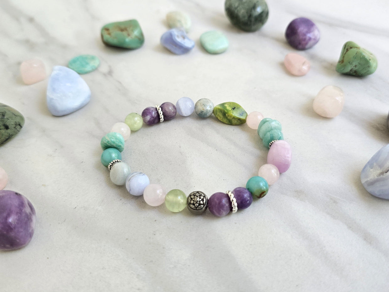 A beaded bracelet with pastel and earthy tones is arranged on a light marble surface, surrounded by polished crystals. Text overlays read 'MOXIE Monthly', 'Joyful Service', and 'September 2026'.
