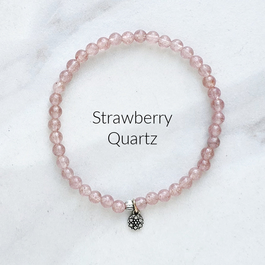 Pink beaded bracelet with a silver charm on a white marble background, labeled 'Strawberry Quartz'.