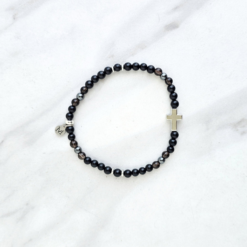 Black beaded bracelet with a cross charm on a white fabric background, the watcher Christian bracelet psalm 121:7-8