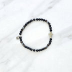 Black beaded bracelet with a cross charm on a white fabric background, the watcher Christian bracelet psalm 121:7-8