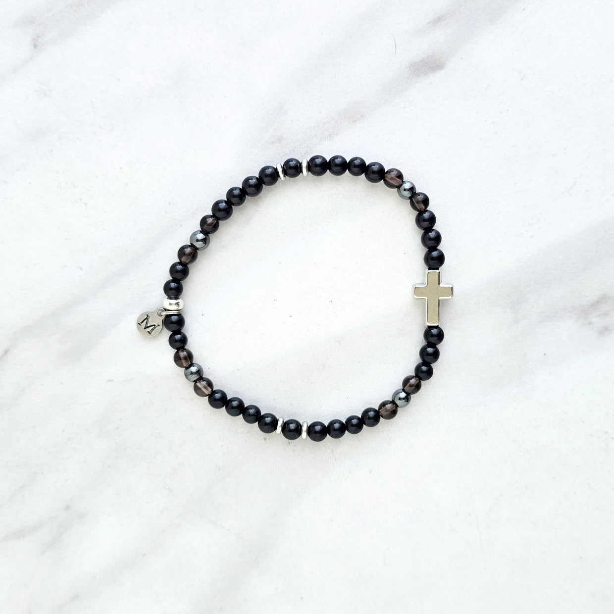 Black beaded bracelet with a cross charm on a white fabric background, the watcher Christian bracelet psalm 121:7-8