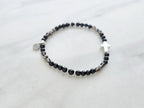 Black beaded bracelet with a cross charm on a white fabric background, the watcher Christian bracelet psalm 121:7-8