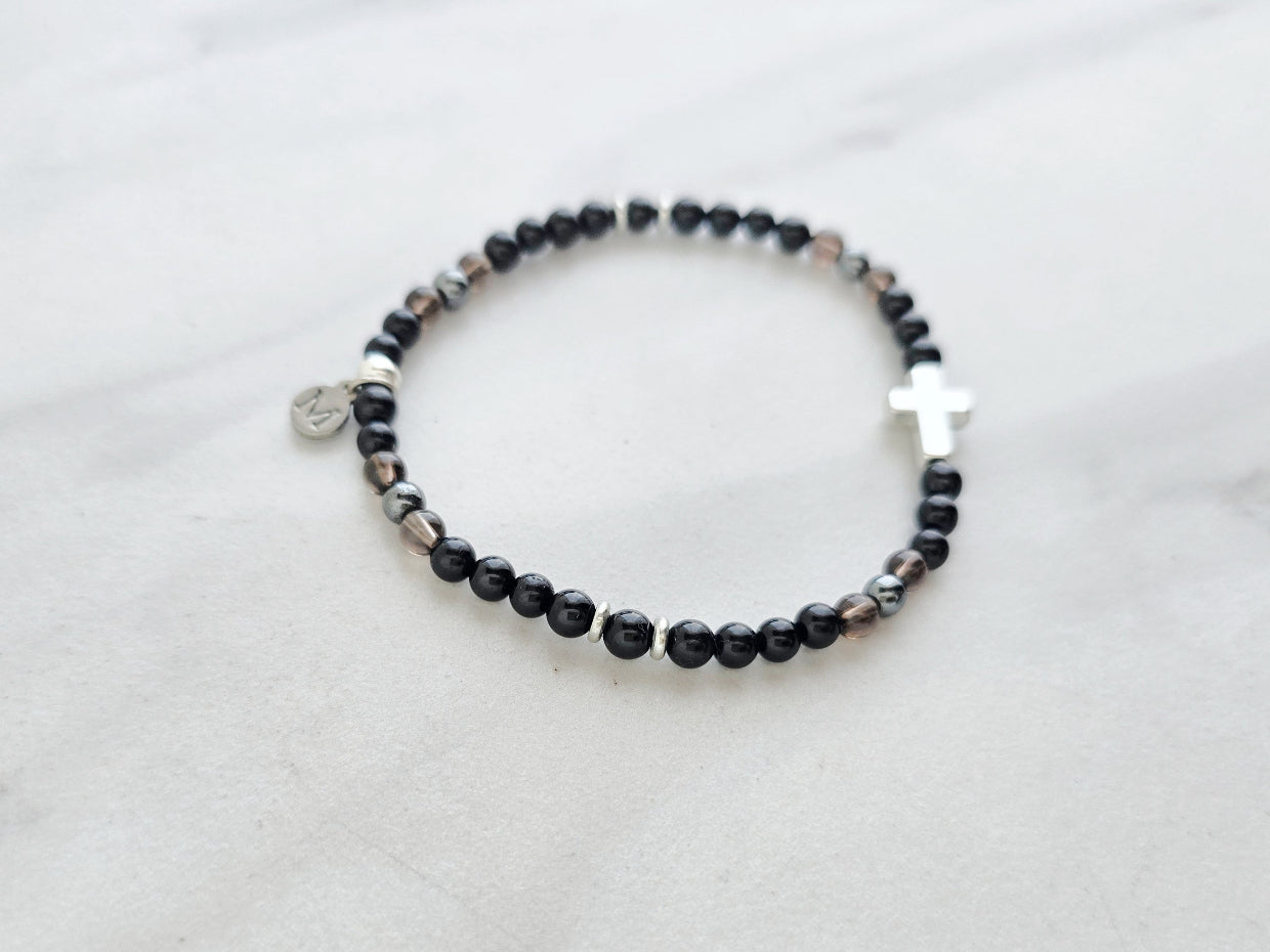 Black beaded bracelet with a cross charm on a white fabric background, the watcher Christian bracelet psalm 121:7-8