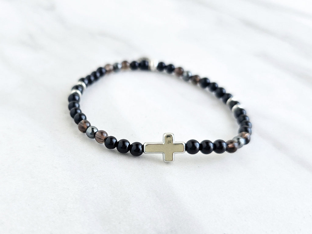 Black beaded bracelet with a cross charm on a white fabric background, the watcher Christian bracelet psalm 121:7-8