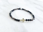 Black beaded bracelet with a cross charm on a white fabric background, the watcher Christian bracelet psalm 121:7-8