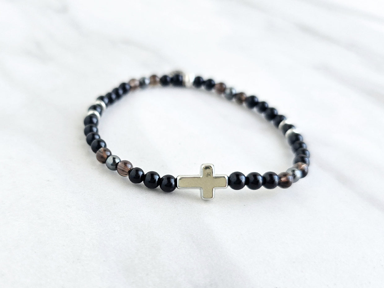 Black beaded bracelet with a cross charm on a white fabric background, the watcher Christian bracelet psalm 121:7-8