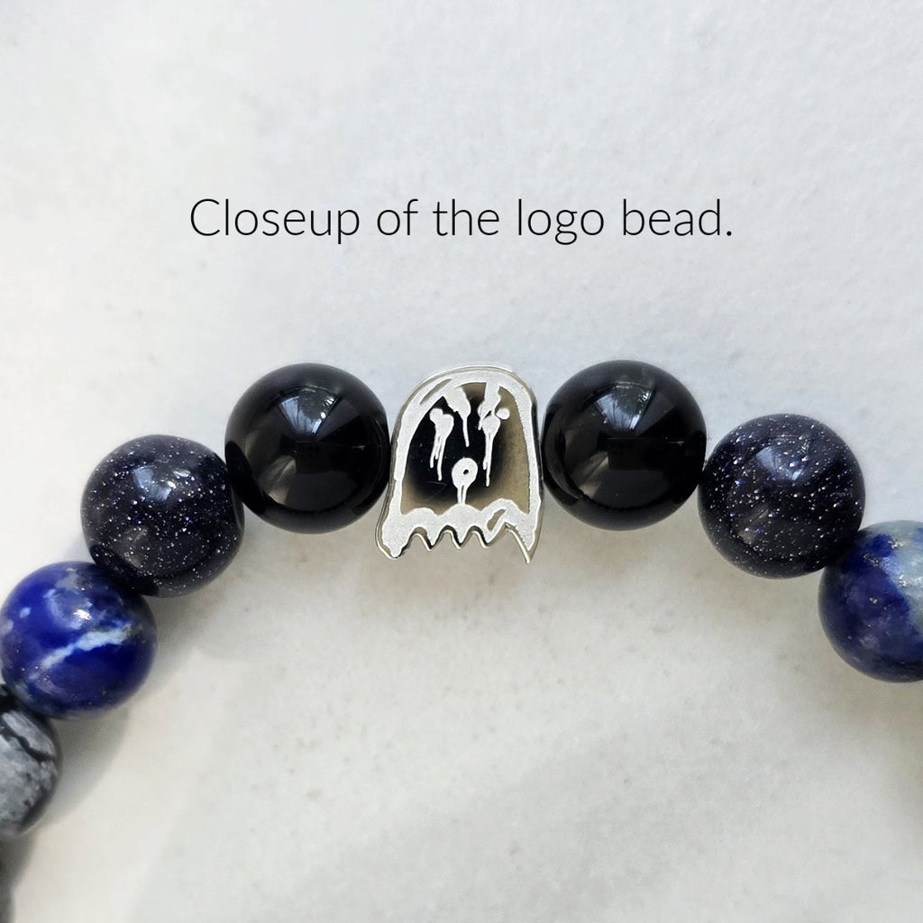 Closeup of a beaded bracelet featuring glossy black and deep blue beads with white speckles, a silver-toned logo charm, and a white background with text reading 'Closeup of the logo bead.'.