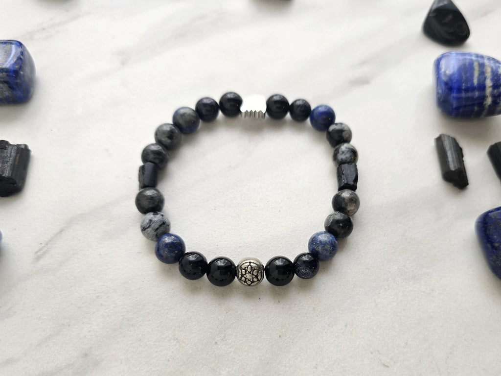 A beaded bracelet with blue, gray, black, and silver beads, displayed on a white marble surface surrounded by black and blue crystals. Text overlays read 'A Collaboration with URBAN INVESTIGATOR + MOXIE MALAS' and 'Abandonment Issues'.