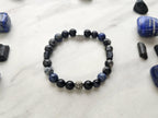 A beaded bracelet with blue, gray, black, and silver beads, displayed on a white marble surface surrounded by black and blue crystals. Text overlays read 'A Collaboration with URBAN INVESTIGATOR + MOXIE MALAS' and 'Abandonment Issues'.