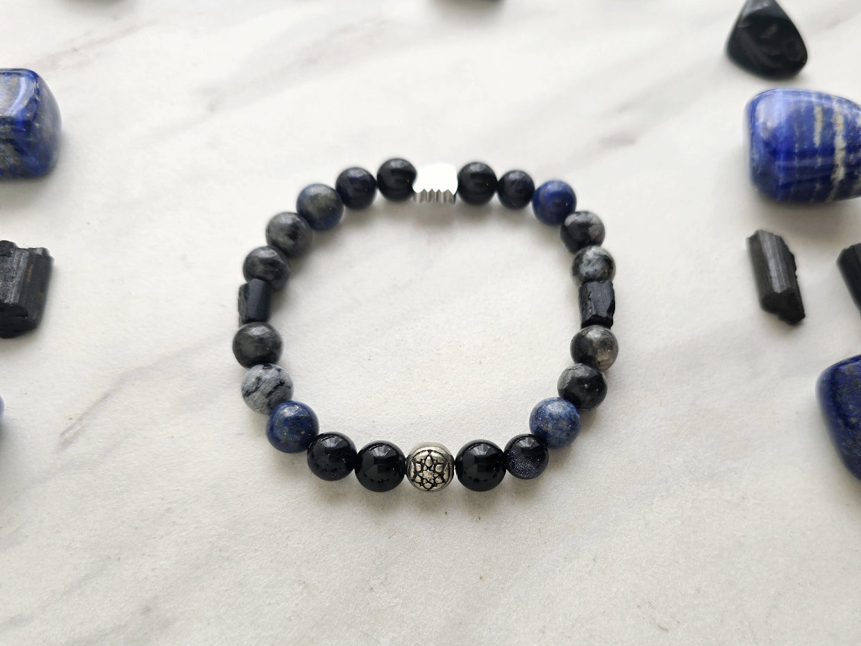 A beaded bracelet with blue, gray, black, and silver beads, displayed on a white marble surface surrounded by black and blue crystals. Text overlays read 'A Collaboration with URBAN INVESTIGATOR + MOXIE MALAS' and 'Abandonment Issues'.