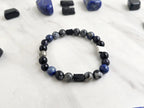 A beaded bracelet with blue, gray, black, and silver beads, displayed on a white marble surface surrounded by black and blue crystals. Text overlays read 'A Collaboration with URBAN INVESTIGATOR + MOXIE MALAS' and 'Abandonment Issues'.