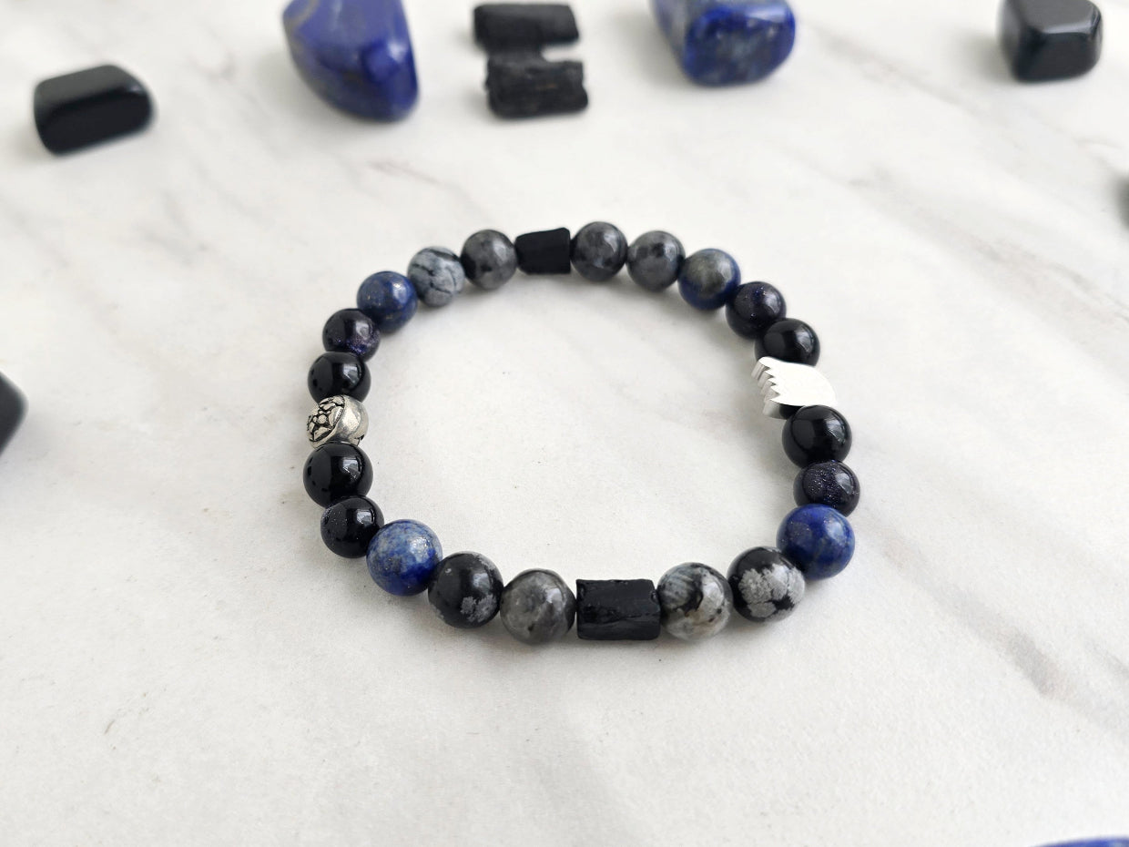 A beaded bracelet with blue, gray, black, and silver beads, displayed on a white marble surface surrounded by black and blue crystals. Text overlays read 'A Collaboration with URBAN INVESTIGATOR + MOXIE MALAS' and 'Abandonment Issues'.
